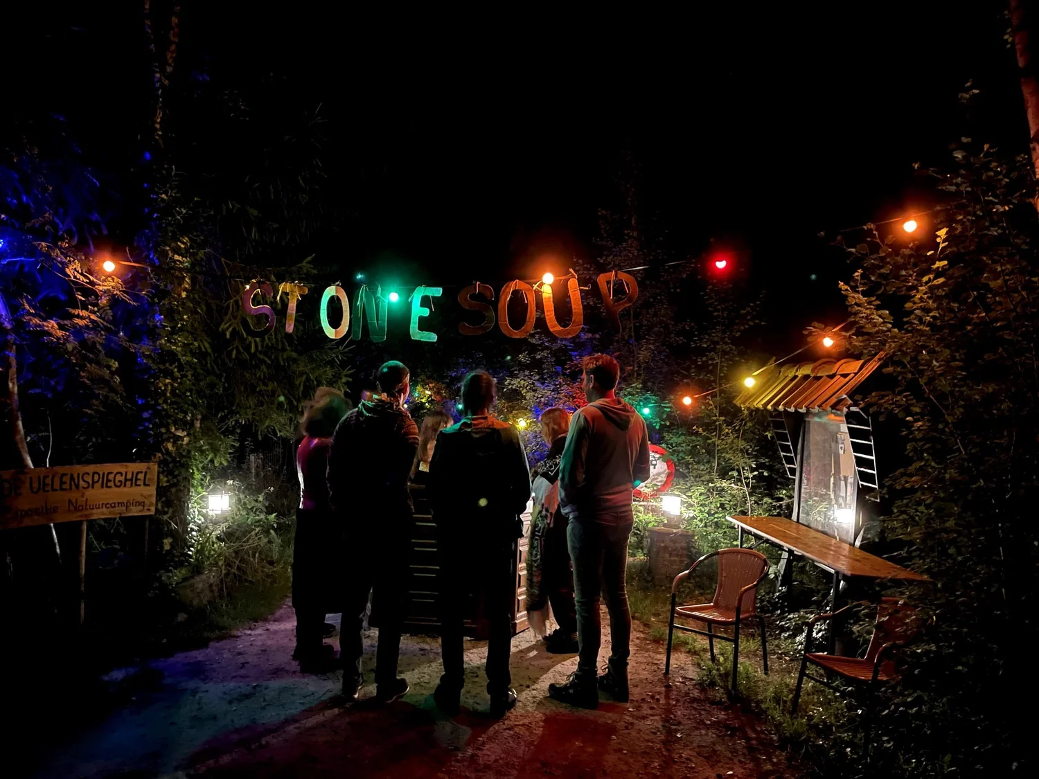 People gathered beneath a handmade Stone Soup sign and glowing lights at night